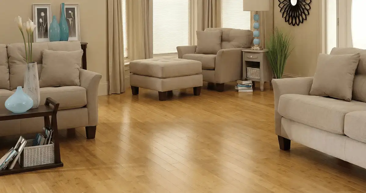 A living room with professionally installed bamboo flooring, beige sofas, a matching ottoman, a side table with a blue lamp, vases, framed pictures, and large windows with blinds and curtains in Riverside, CA