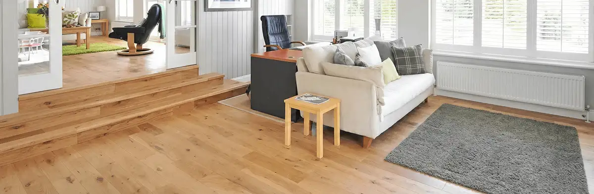 A modern living room with professionally installed laminate flooring, a beige sofa, a small side table, a gray rug, and large windows with white shutters in Riverside, CA